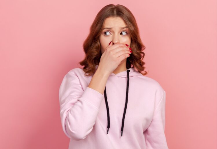 Portrait of shocked young curly haired teen girl in casual hoodie closing mouth with hand, keeping secret information, oh my god gesture. Indoor studio shot isolated on pink background